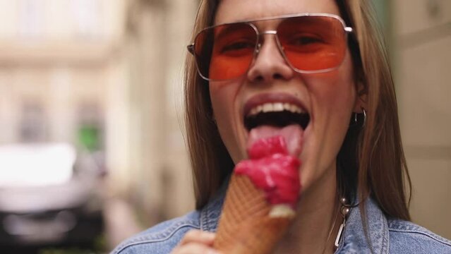 Close up happy young woman with delicious pink ice cream in waffle cone outdoors. Girl wear orange sunglasses. Blonde female lick ice cream and laughing, she enjoy. Slow motion.