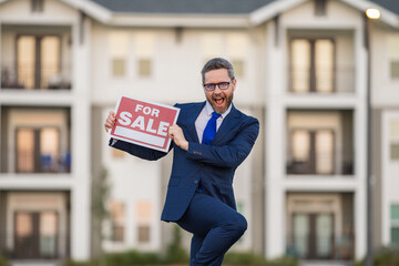 Funny realtor agent is a realtor with sign for sale in hand against the background on new apartment home background. Realtor in suit, outdoor portrait. Realtor offering new home. Property concept.