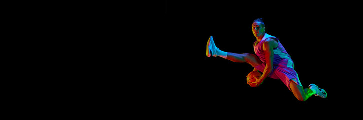 Obraz premium Dynamic image of teen boy during basketball game in motion, jumping with ball against black background in neon light. Concept of professional sport, competition, competition. Banner. Copy space for ad