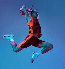 Scoring winning goal. Young man in uniform playing basketball, jumping with ball against blue studio background in neon light. Concept of professional sport, competition, hobby, game, competition © master1305