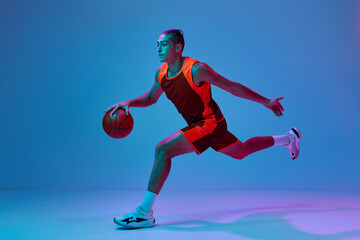 Fototapeta na wymiar Dynamic image of teen boy in uniform, playing basketball against blue studio background in neon light. Dribbling ball. Concept of professional sport, competition, hobby, game, competition