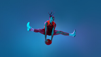 Twine in air. Sportive young guy, basketball player in motion during match, jumping with ball over...