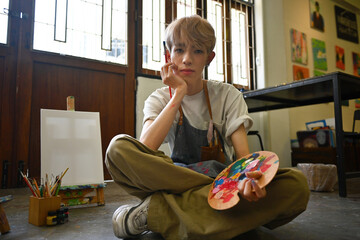 Portrait image of LGBT artist, an Asian male teenager with colored hair, looking at the camera with confidence while sitting on the floor in the workshop studio