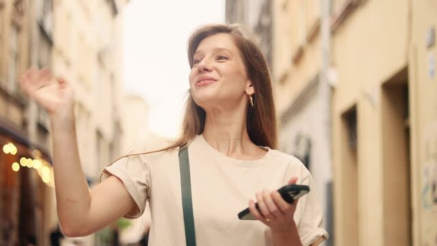 Pretty young woman hold smartphone scrolling social media texting browsing online walking down street in city centre while meet and wave to someone Positive female relax enjoying great day outdoors 