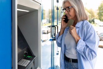 mature gray-haired woman standing at an ATM calling a hotline