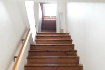 Un ancien escalier avec des marches en bois et une rambarde en bois, face à une porte ouverte qui baigne dans lumière