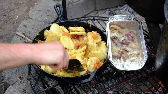 Stirring almost tender potatoes slices and carrots while being cook in cast iron pan over fire pit alongside duck meat odds like neck and gizzard in the aluminum tray.