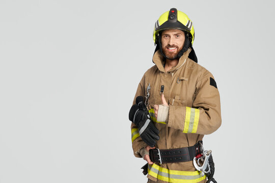 Happy caucasian firefighter in uniform showing thumb up in studio. Front view of confident bearded fireman giving like, while smiling to camera, on gray background. Concept of work, hand gesture.