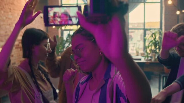 Selective focus slowmo portrait of attractive young African American woman filming while dancing with her friends at party in loft studio, 90s aesthetics