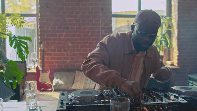 Portrait of joyful young African American DJ playing music set using mixer console and dancing during party in loft room