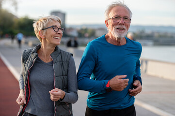 Cheerful active senior couple jogging together outdoors along the river. Healthy activities for...