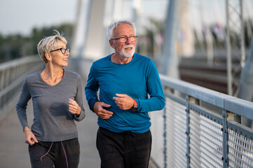 Cheerful active senior couple jogging together outdoors on the bridge. Healthy activities for...