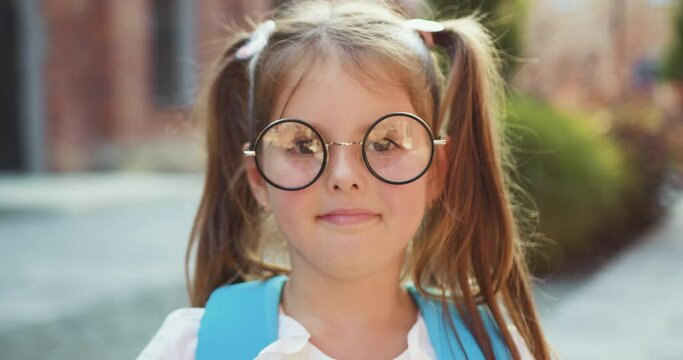 Close up. Portrait of lovely small school girl in glasses with backpack smiling posing on camera near campus. Female elementary school student holds textbooks standing outside school building.