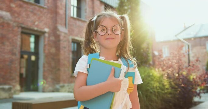 Portrait little girl in glassas with backpack and books in her hand on a school building background. Happy school girl smiling at camera. Child goes to first grade. Education concept.