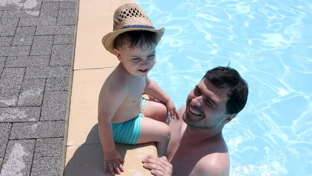 kid on poolside.sibling playing with legs in pool water,summer vacation time. dad kissing little child.baby boy in beach hat 4k 