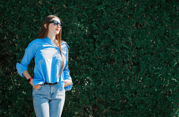 Fototapeta premium Summer portrait of a girl with long blond hair in sunglasses. A girl in a blue shirt and blue jeans stands against a background of greenery.