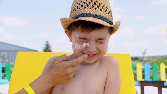 boy kid sitting on beach pool lounger with hat on head.mother painting sun shape from sunblock cream lotion on face cheek. hand applying lotion on shoulder,back 4k summer vacation time