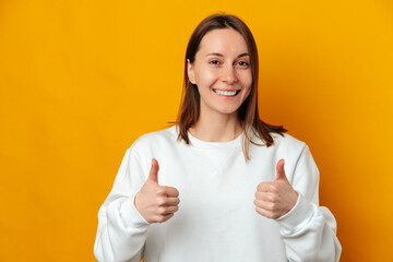 Handsome young smiling woman wearing white shirt is showing thumbs up over yellow background.