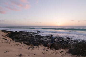 person looking at the waves of the sea sitting on a rock, while the sun rises.