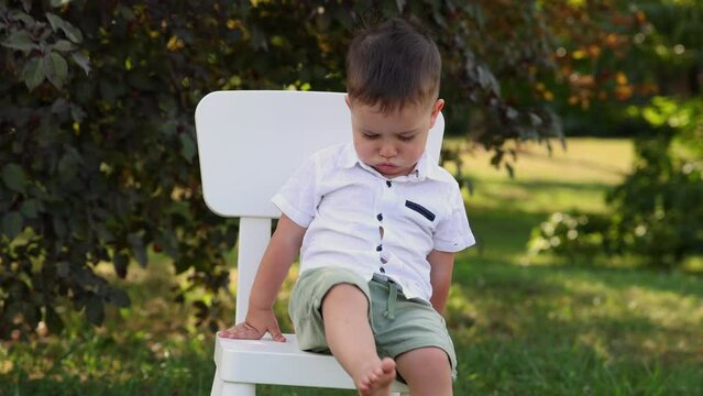 upset cute baby boy in park celebrating birthday sitting on chair,eating mini cake.kid hit the balloon with feet. angry dissatisfied child making funny face expression,carrying chair with him.