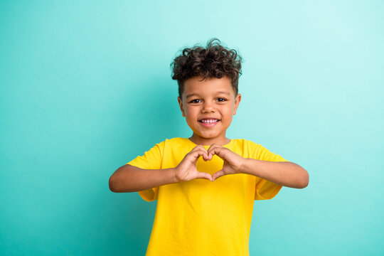 Portrait of appreciative small boy with curly hair wear stylish t-shirt fingers show heart symbol isolated on turquoise color background