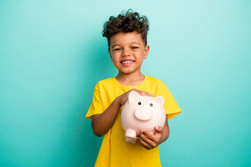 Portrait of toothy beaming small boy with brown hair wear stylish t-shirt arms holding piggy box isolated on teal color background © deagreez