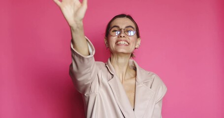 Happy friendly young woman waves palm in hello gesture welcomes someone with hospitable expression, positive emotions dressed casually isolated over pink background. Hi to all of you.