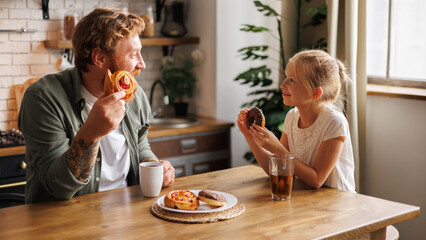 Smiling girl holding donut and looking at tattooed father during breakfast together at home