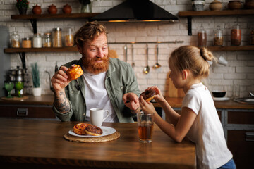 Cheerful tattooed father holding pastry and pointing at daughter while talking during breakfast in kitchen 