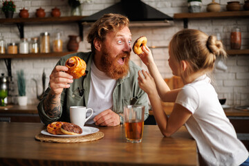 Father having fun with daughter and pastry during breakfast in kitchen 