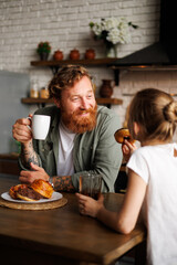Cheerful bearded father holding coffee while daughter eating donut during breakfast in kitchen 