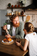 Bearded and tattooed father holding coffee and looking at blurred daughter with donut during breakfast at home