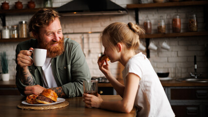 Smiling tattooed father holding coffee while daughter eating donut in morning in kitchen 