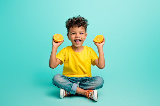 Full length photo of good mood small boy with brown hair wear stylish t-shirt sit hold sliced orange isolated on teal color background - Powered by Adobe