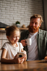 Cheerful freckled kid using smartphone near serious father with coffee in kitchen at home, internet addiction concept