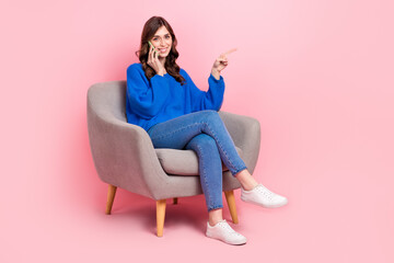 Obraz na płótnie Canvas Full size photo of charming cheerful girl sit soft chair speak telephone indicate finger empty space isolated on pink color background