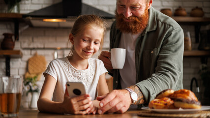 Smiling bearded father holding coffee and talking to daughter with smartphone during breakfast in kitchen 