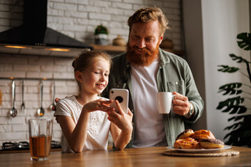 Fototapeta na wymiar Smiling freckled child showing smartphone to bearded father with coffee in kitchen during breakfast 