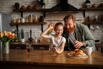 Bearded parent and cheerful daughter using smartphone together near coffee and sweet pastry in kitchen 