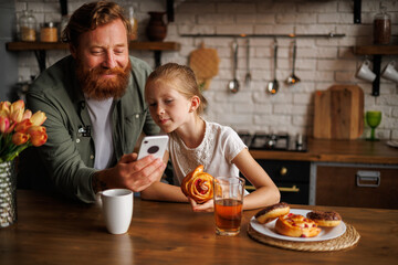 Smiling father using smartphone with daughter holding pastry during breakfast at home