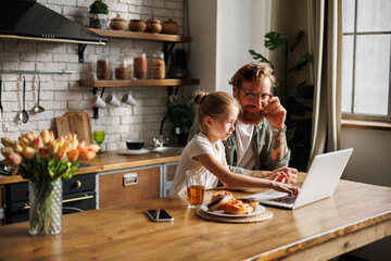 Child using laptop together with tattooed father during breakfast near pastry in kitchen 