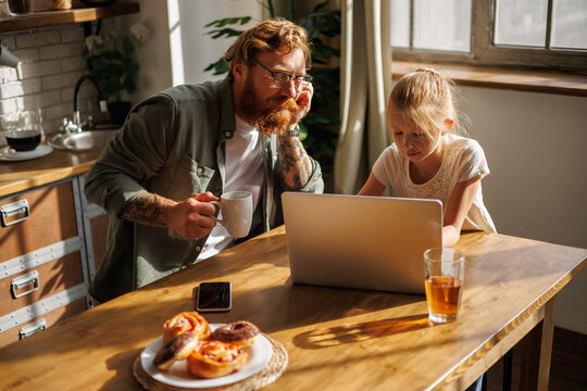 Tattooed father holding coffee and using laptop with daughter near pastry in kitchen in morning