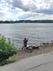 A fisherman stands on the bank of a wide river and catches fish with a fishing rod. Rear view, face unknown