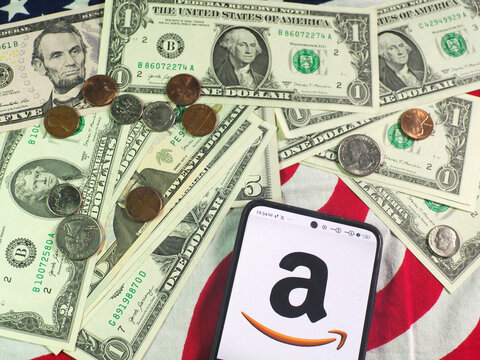 In this photo illustration,  Amazon.com, Inc. logo is seen displayed on a smartphone and US currency notes and coins in the background.