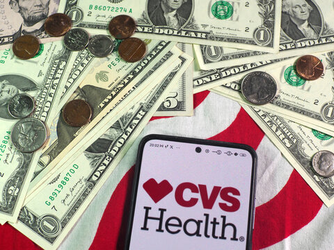 In this photo illustration,  CVS Health Payor Solutions logo is seen displayed on a smartphone and US currency notes and coins in the background.
