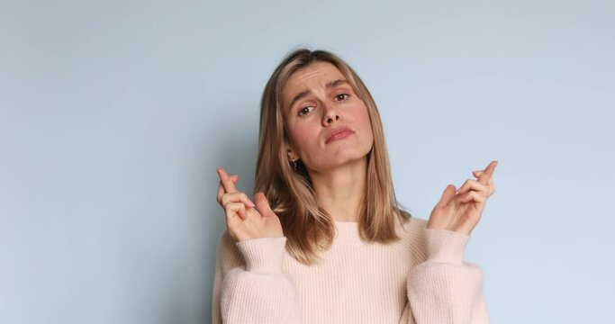 Pleading begging young blonde girl wears ecru sweater hold hands folded in prayer making wish keep fingers crossed isolated on pastel light blue background. People emotions lifestyle concept.