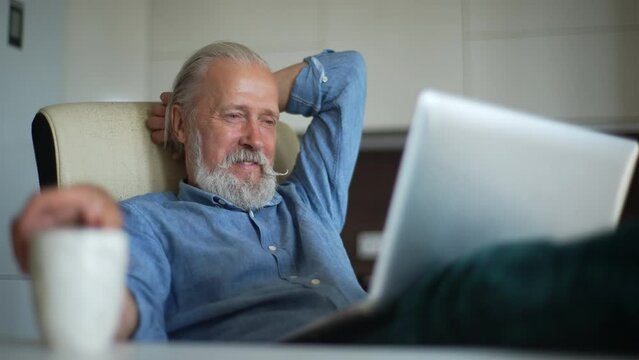 Happy senior aged man sitting in comfort with coffee cup working with laptop computer in home office. Relaxed mature adult male drinking coffee using laptop sitting on table at living room.