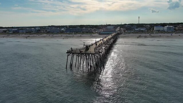 orbit aerial footage of Kure Pier with people fishing, ocean water, waves and condominiums and homes along the coast with blue sky and clouds at sunset in Wilmington North Carolina USA