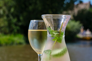 A glass of white wine and a bottle of water with mint leaves and a slice of lime on a sunny terrace by the water. Image with selective focus.