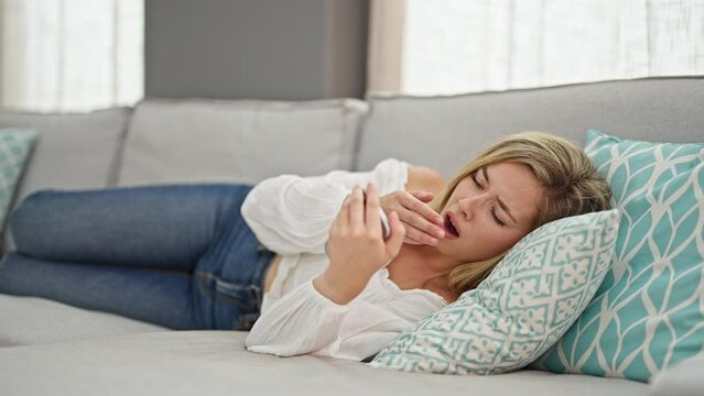 Young blonde woman using smartphone lying on sofa tired sleeping at home